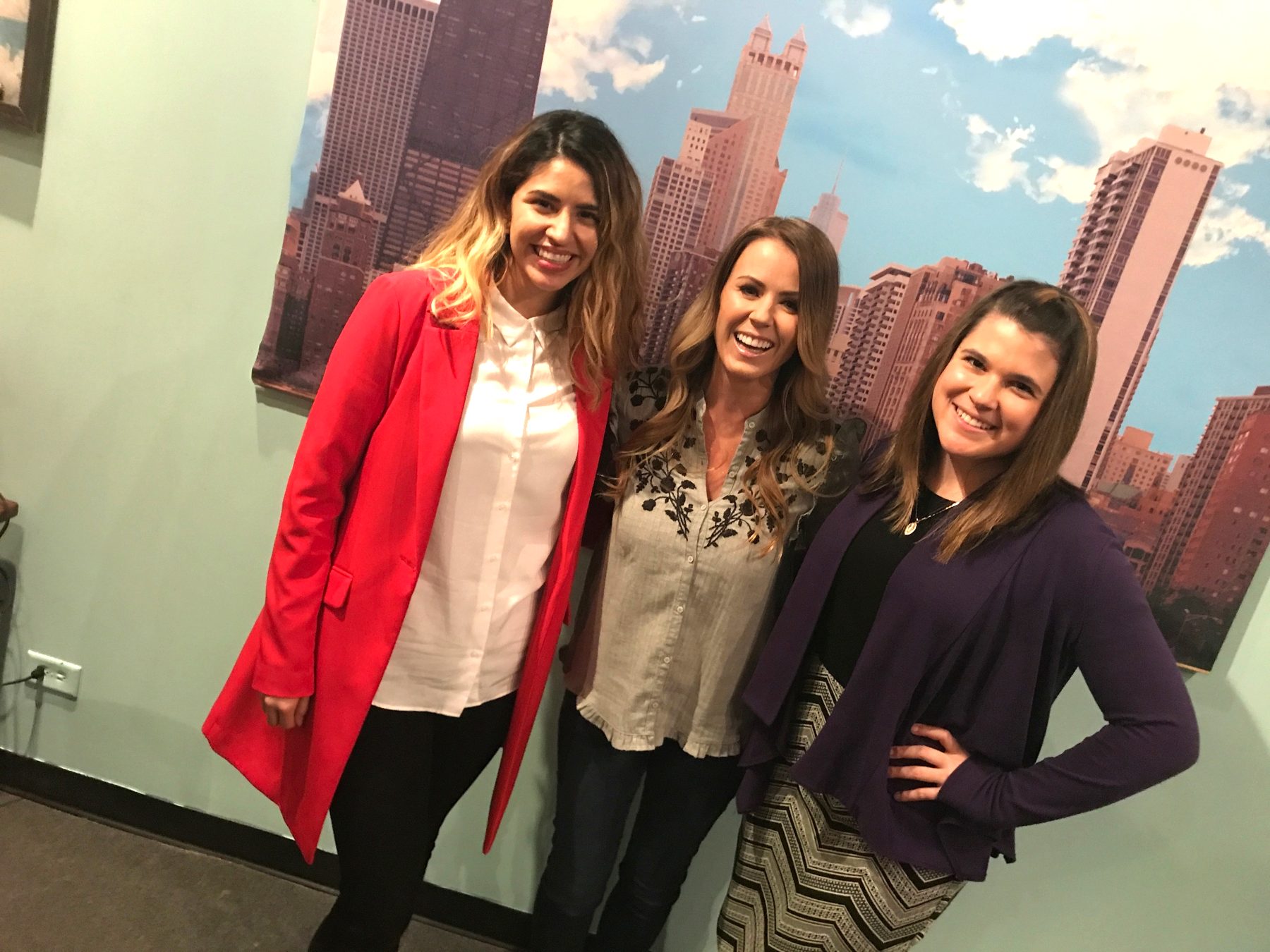 Three smiling women posing together in front of a printed city-skyline backdrop