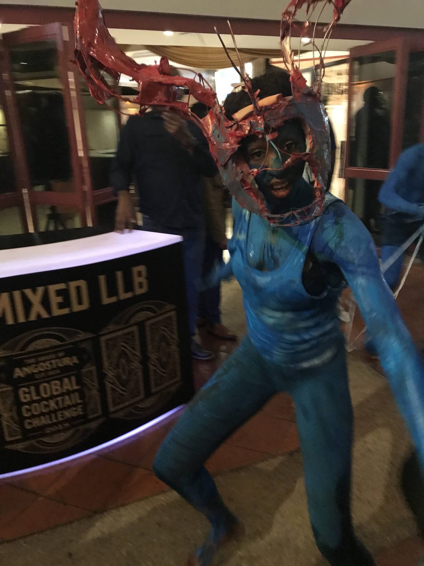 A carnival performer in blue body paint and a horned mask beside a Global Cocktail Challenge bar
