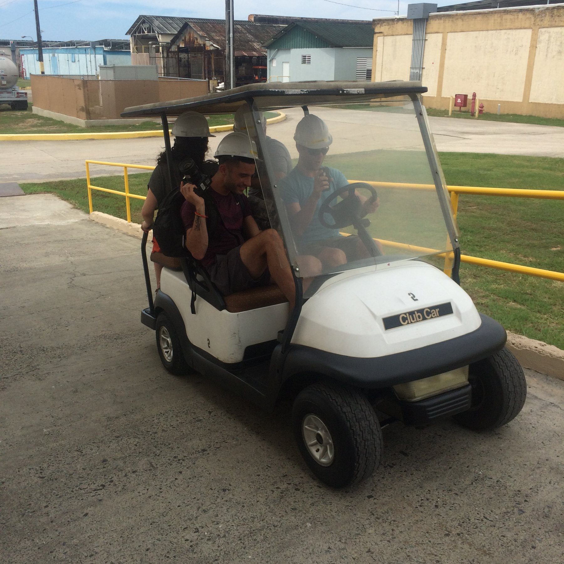Journalists in hard hats, one holding a camera, riding a golf cart through a distillery campus