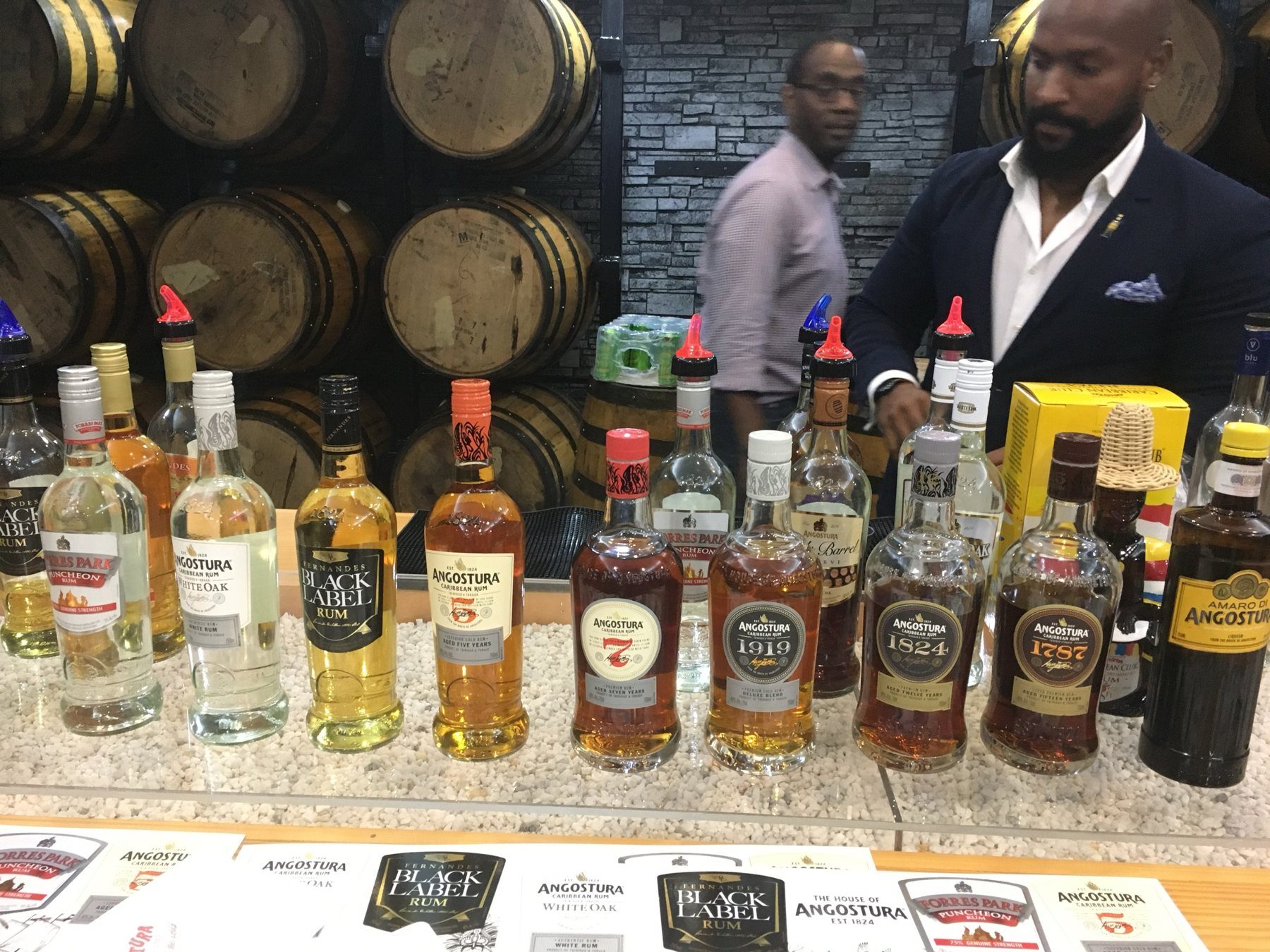 A row of Angostura rums including the 5, 7, 1919, 1824 and 1787 on a bar in front of stacked barrels