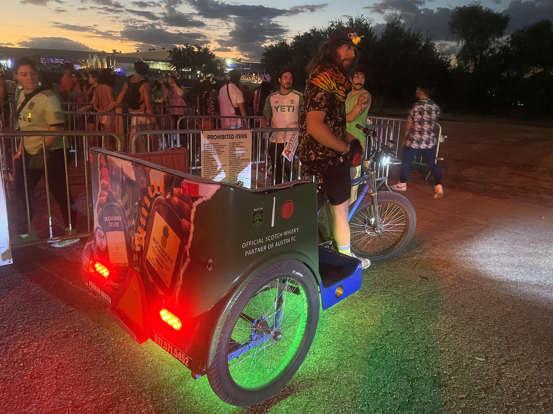 A pedicab at an evening event with panels reading 'Official Scotch Whisky Partner of Austin FC' and Buchanan's DeLuxe and Pineapple bottle art