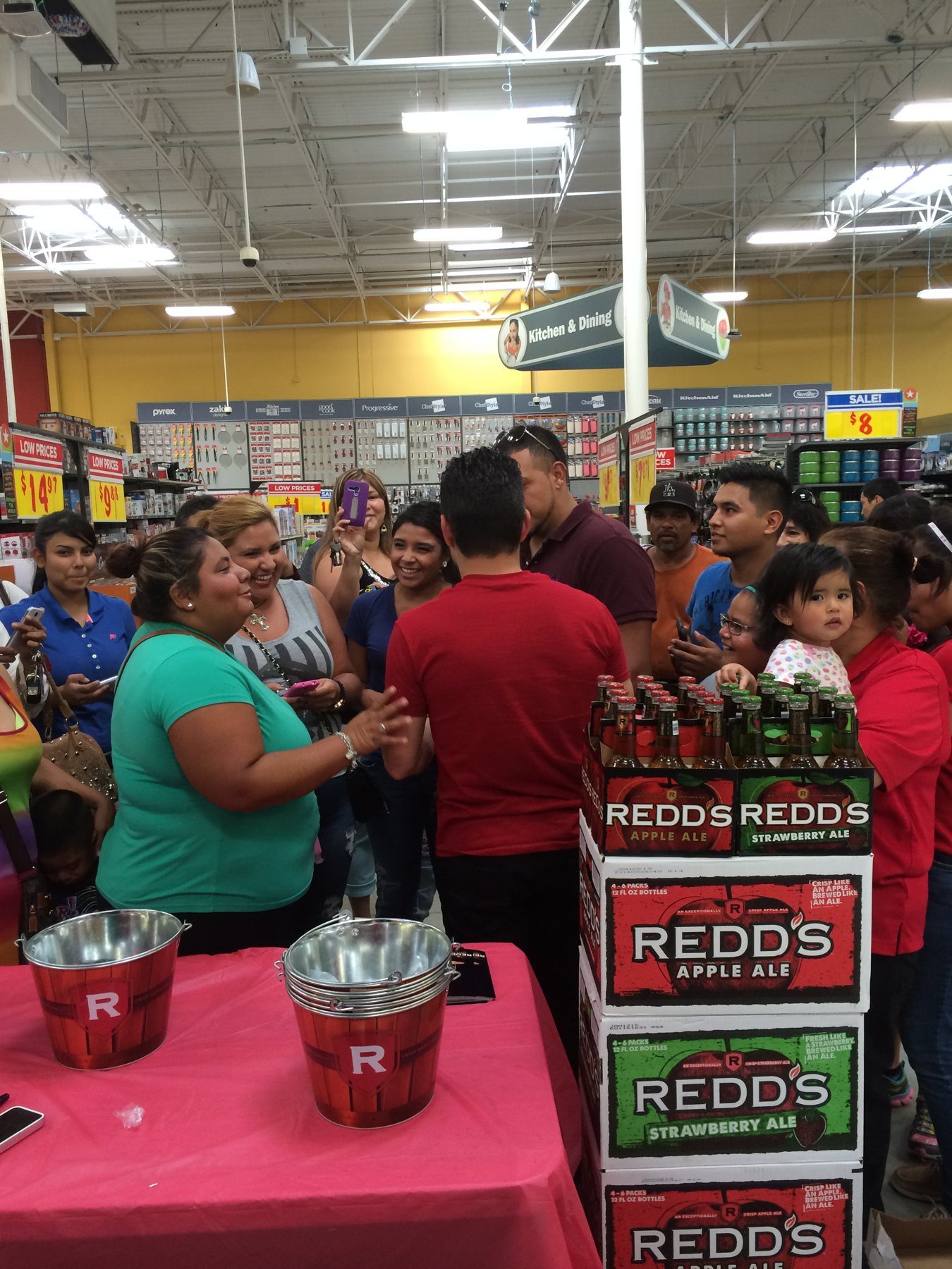 A crowd of shoppers gathered around a guest beside a stacked Redd's Apple Ale and Strawberry Ale display