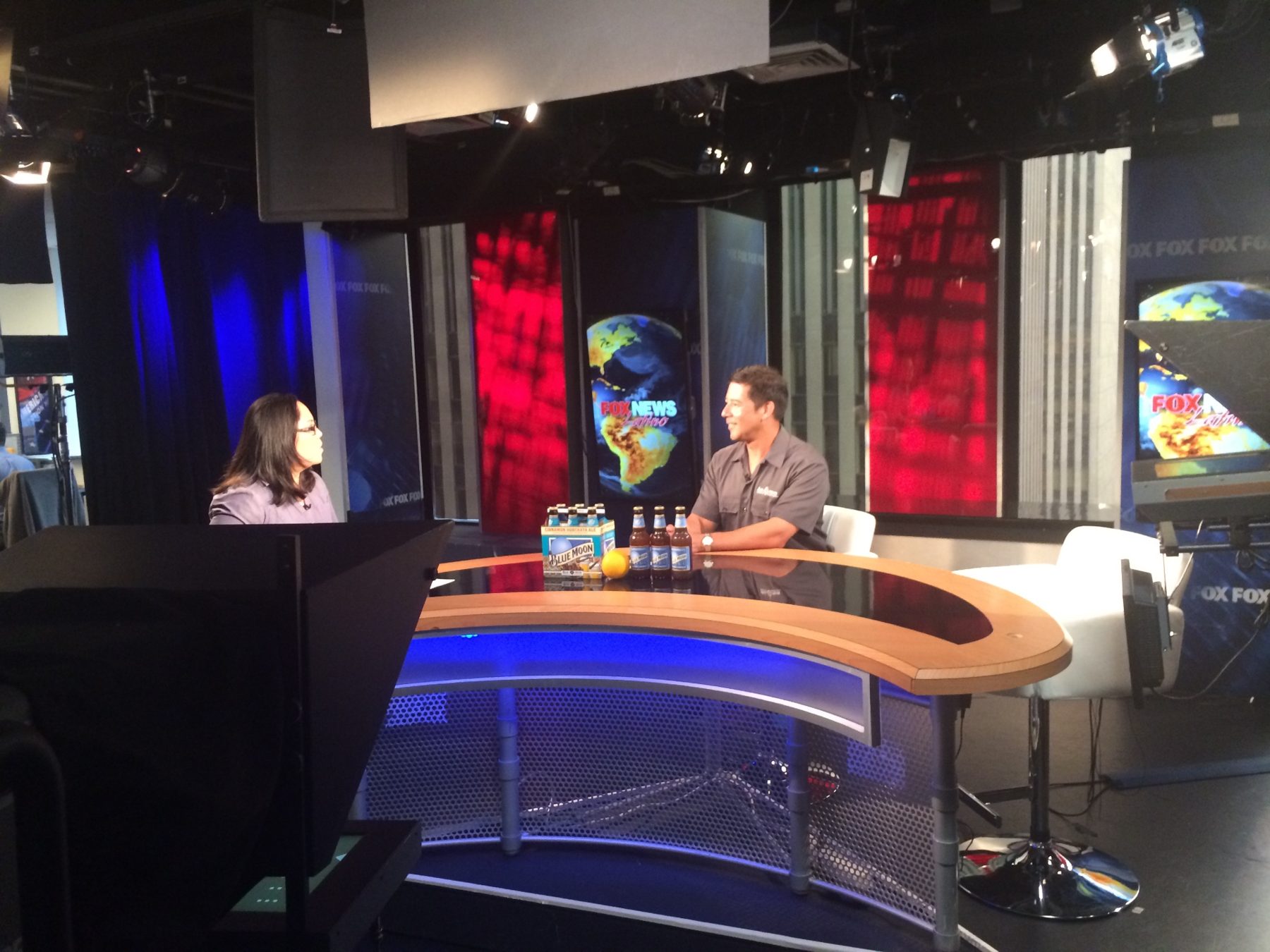 A host interviewing a guest at a Fox News Latino studio desk with Blue Moon bottles and an orange on display