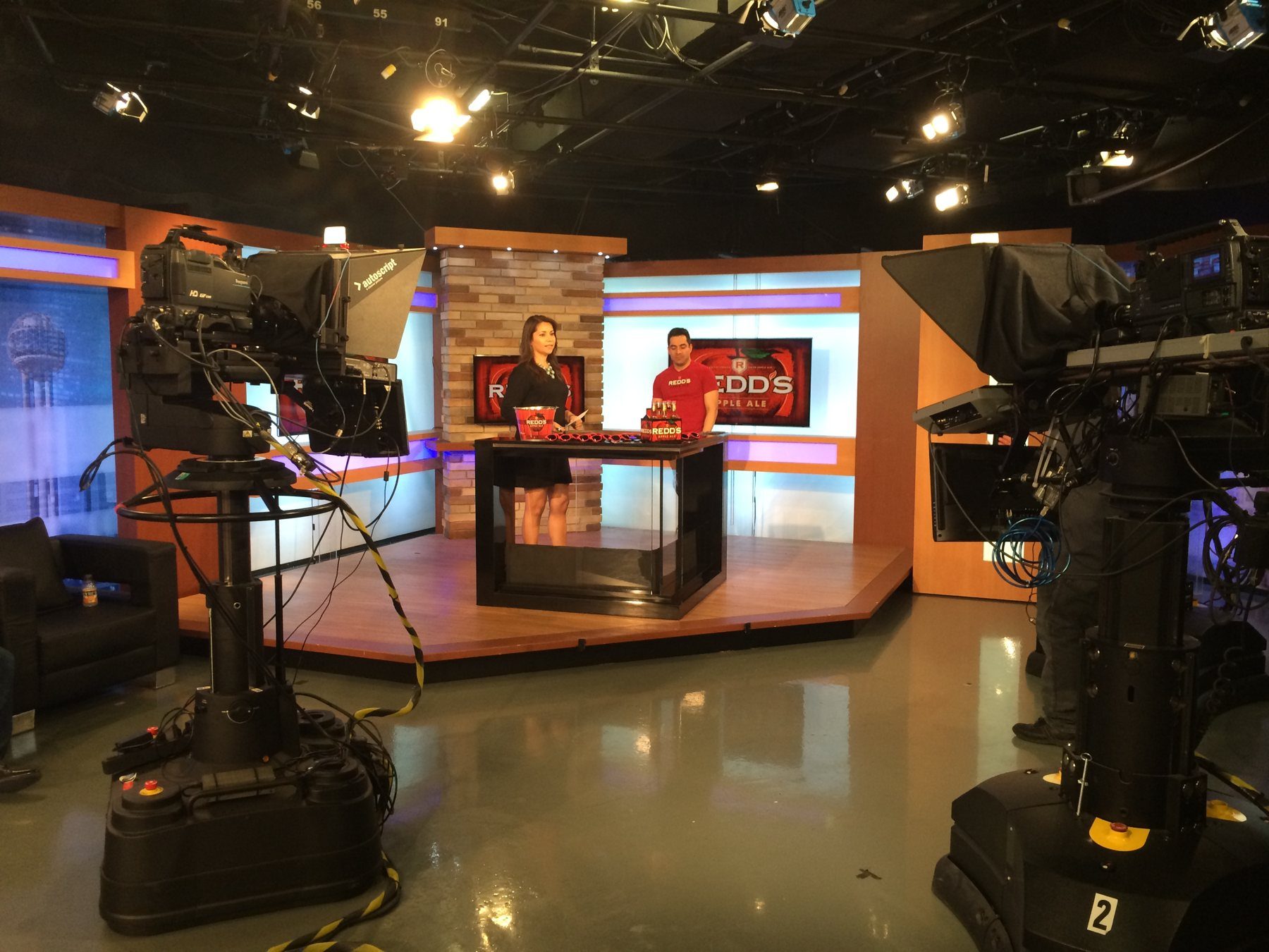 A TV host and a guest in a Redd's Apple Ale shirt on a studio set with Redd's branding and broadcast cameras in the foreground