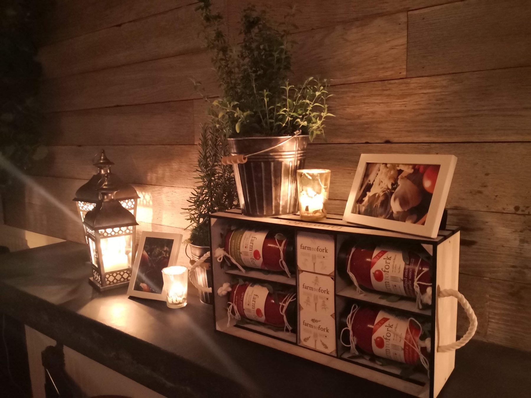A wooden crate holding FarmToFork pasta sauce jars beside framed photos, potted herbs and lanterns lit by candles