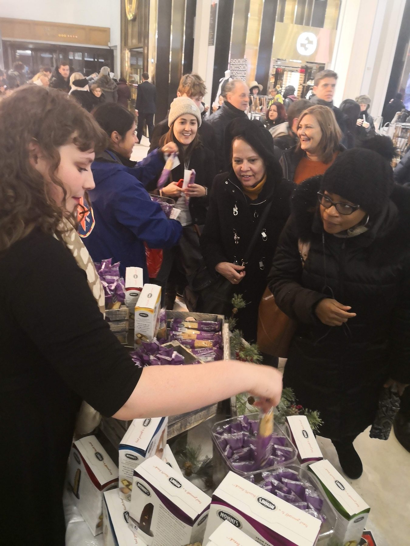 Guests in winter coats gathered around a Nonni’s sampling table stacked with biscotti boxes and purple-wrapped samples