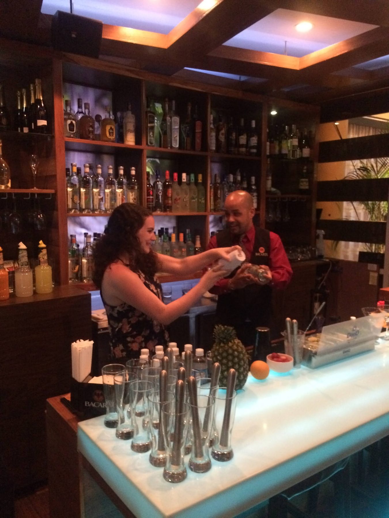 A journalist mixes a cocktail with a bartender behind a lit bar, with a pineapple and fresh fruit on the counter